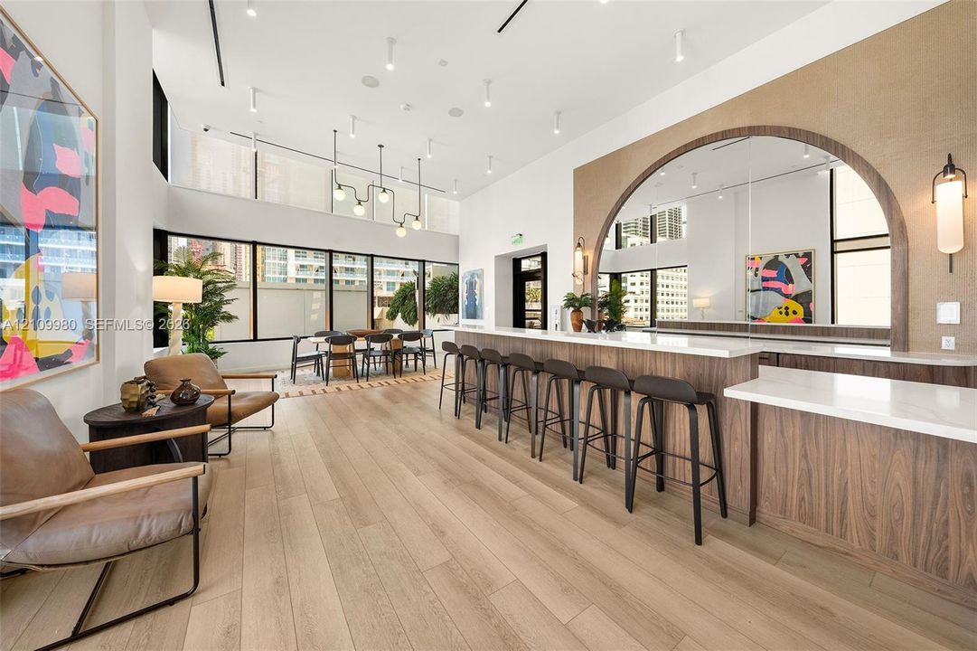 High-ceiling interior featuring wood-finish flooring, a large bar with a stone-surface countertop, an arched mirror display, and expansive floor-to-ceiling windows