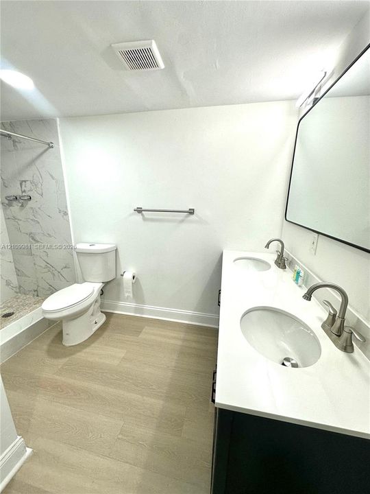 Dual-sink vanity with a solid-surface countertop, wood-finish flooring, and a walk-in shower featuring large-format tile surround
