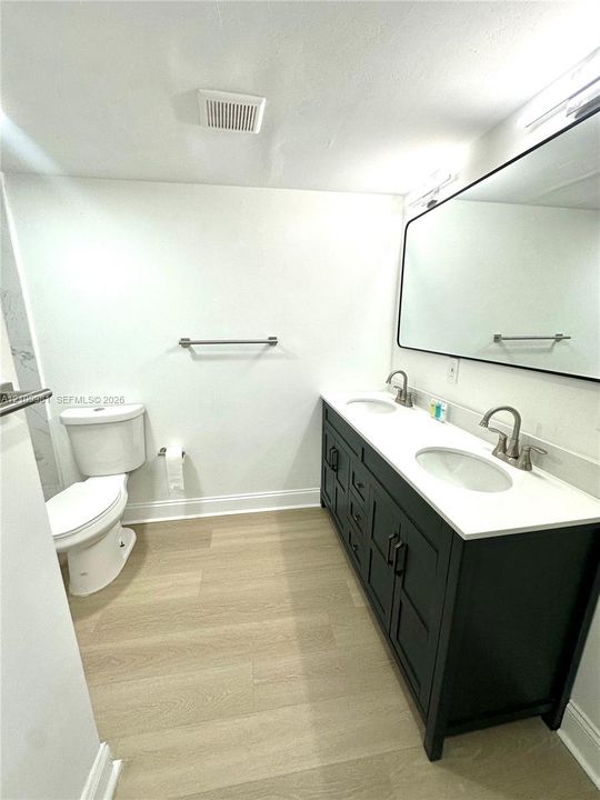 Dual-sink vanity with a dark-finished cabinet, white stone-composite countertop, and metal faucets, paired with a large rectangular mirror and light wood-finish flooring