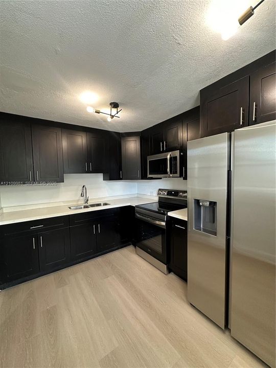 Kitchen featuring dark cabinetry, stainless steel appliances, light-toned countertops, wood-finish flooring, and multi-light ceiling fixture