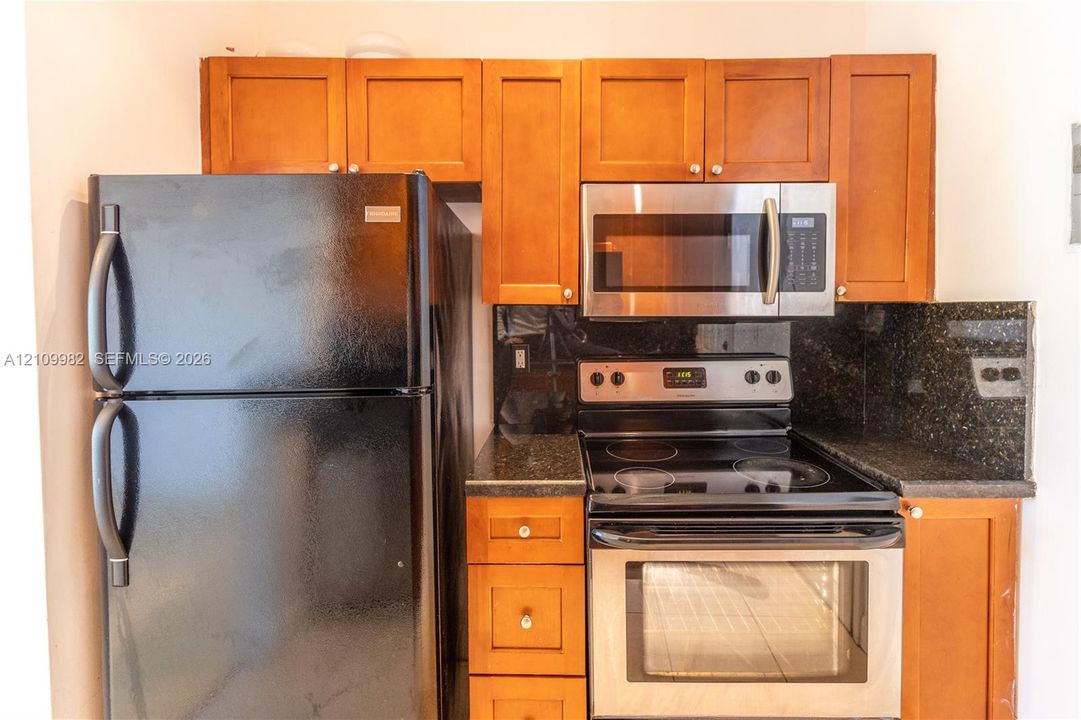 Kitchen featuring shaker-style wood cabinetry, dark stone-finish countertops, stainless steel microwave, glass-top range, and black refrigerator