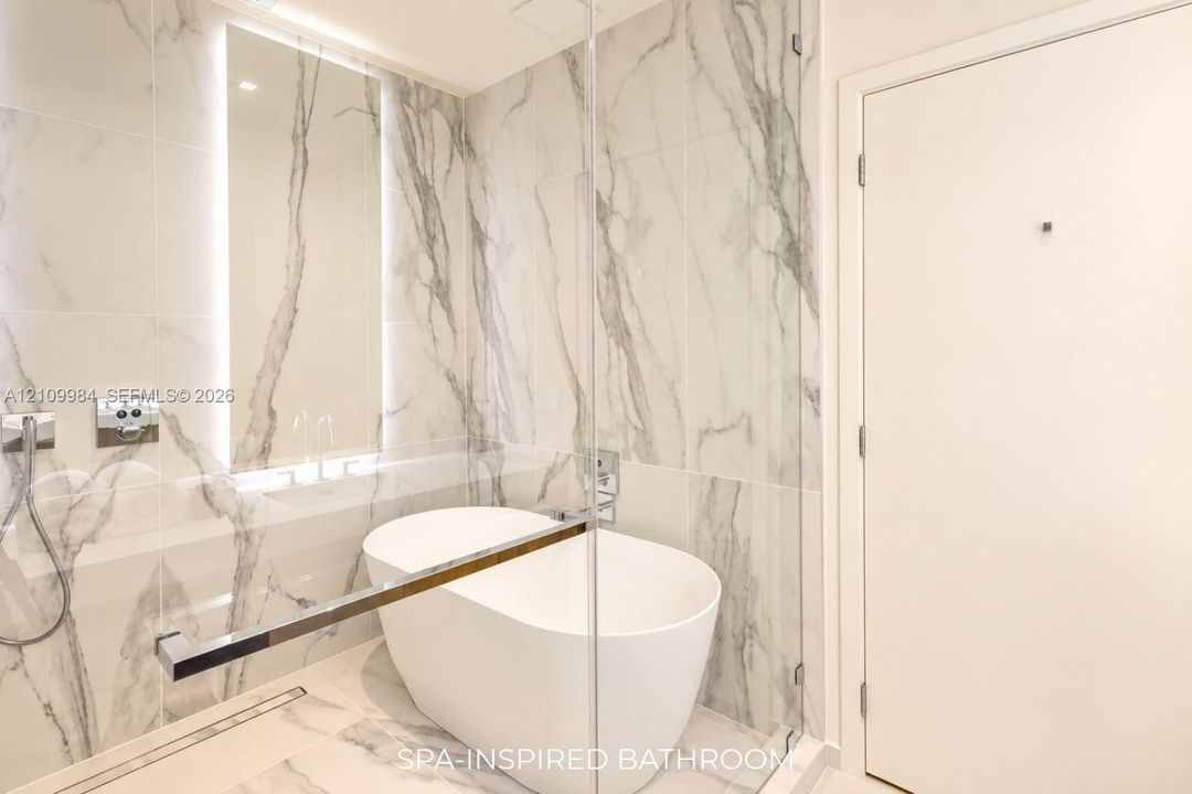 Freestanding soaking tub enclosed in a glass-paneled wet room with floor-to-ceiling stone-patterned tile, integrated wall lighting, and chrome fixtures