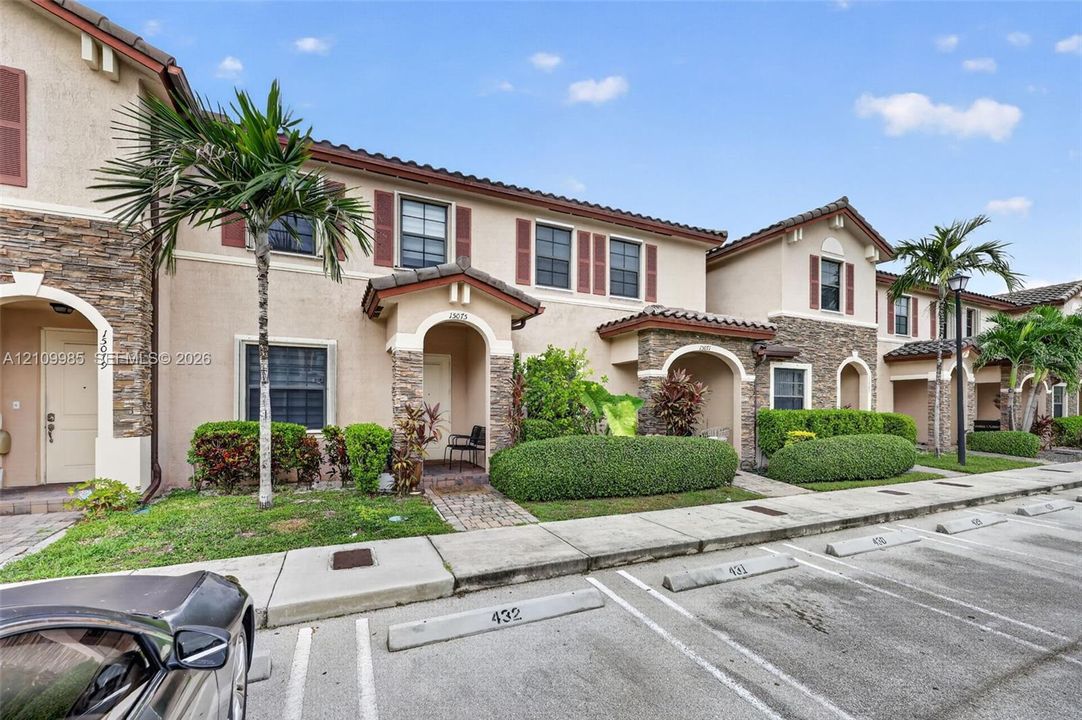 Mediterranean-inspired townhouse exterior featuring stone veneer accents, stucco facade, decorative shutters, and tile roofing, complemented by manicured shrubbery and mature palm trees