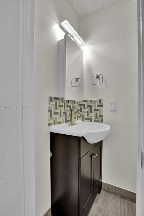 Bathroom vanity featuring a dark wood-finish cabinet, white ceramic sink, herringbone-patterned tile backsplash, wall-mounted medicine cabinet, and linear light fixture