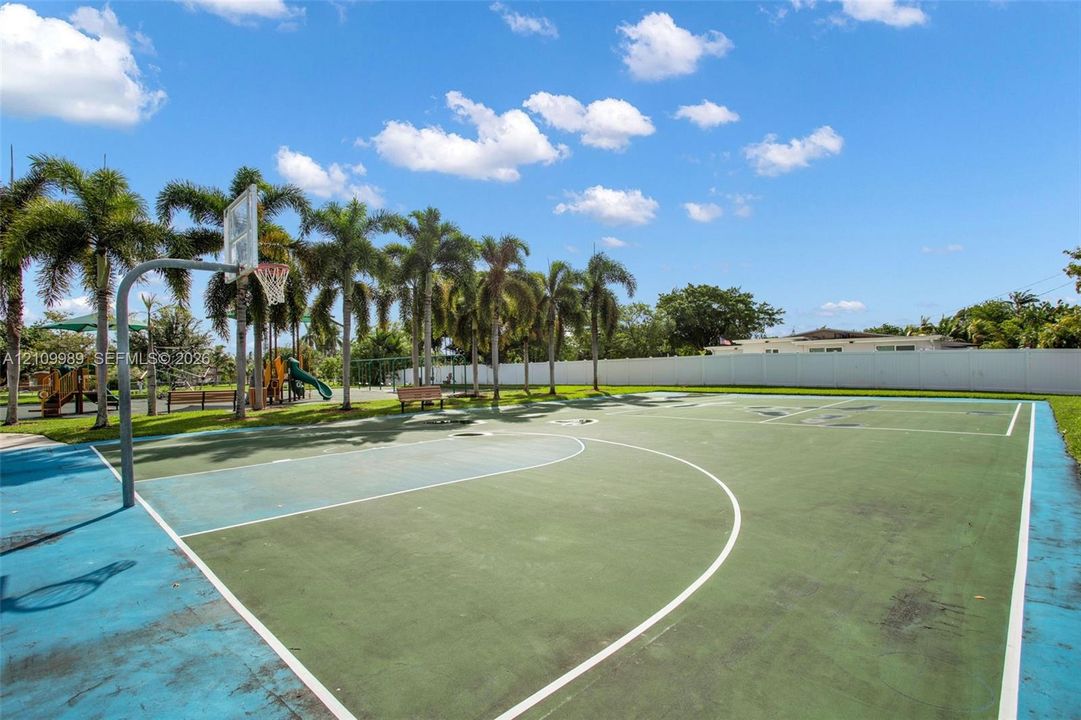 Outdoor basketball court featuring a multi-colored surface, integrated hoop, perimeter fencing, and mature palm trees