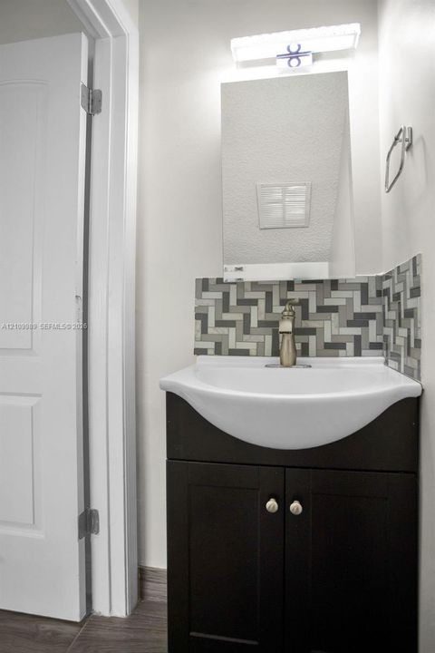 Dark wood-finish vanity with a white integrated basin, multi-tonal herringbone tile backsplash, metallic faucet, wall-mounted rectangular mirror, and bar-style light fixture