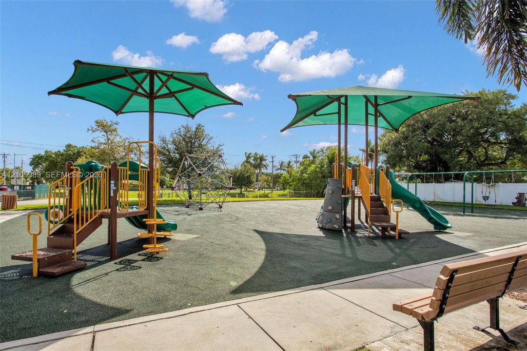 Outdoor recreational area featuring shaded play structures, climbing elements, slides, a swing set, and paved walkways