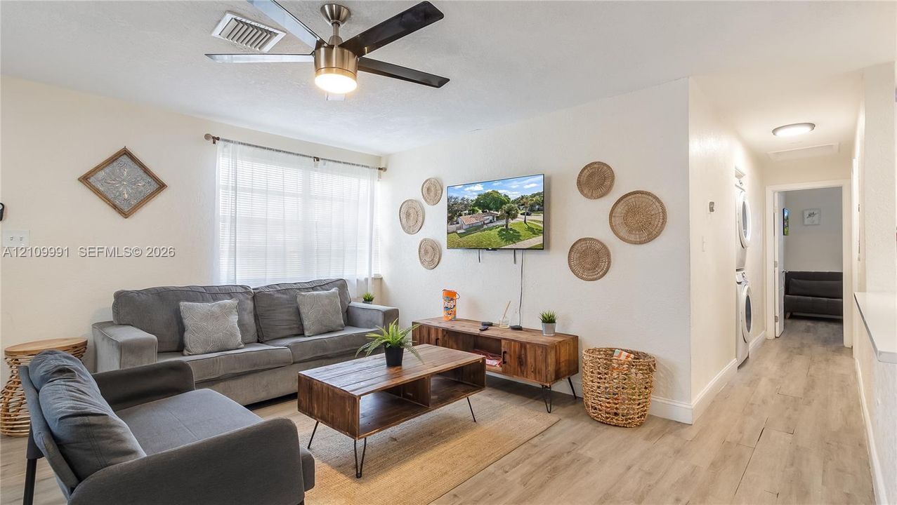Living area featuring wood-finish flooring, a multi-blade ceiling fan with integrated lighting, and a stackable laundry unit