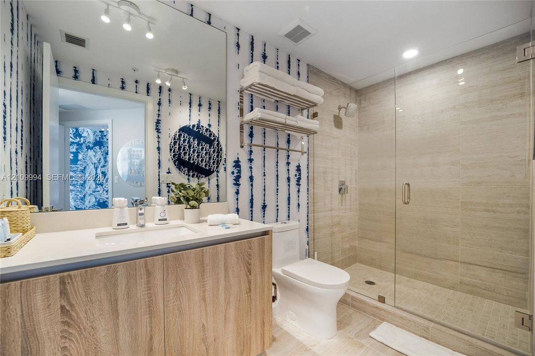 Bathroom featuring a wood-finish vanity with an integrated countertop, a glass-enclosed walk-in shower with textured tile surround, and a wall-mounted stainless steel towel rack