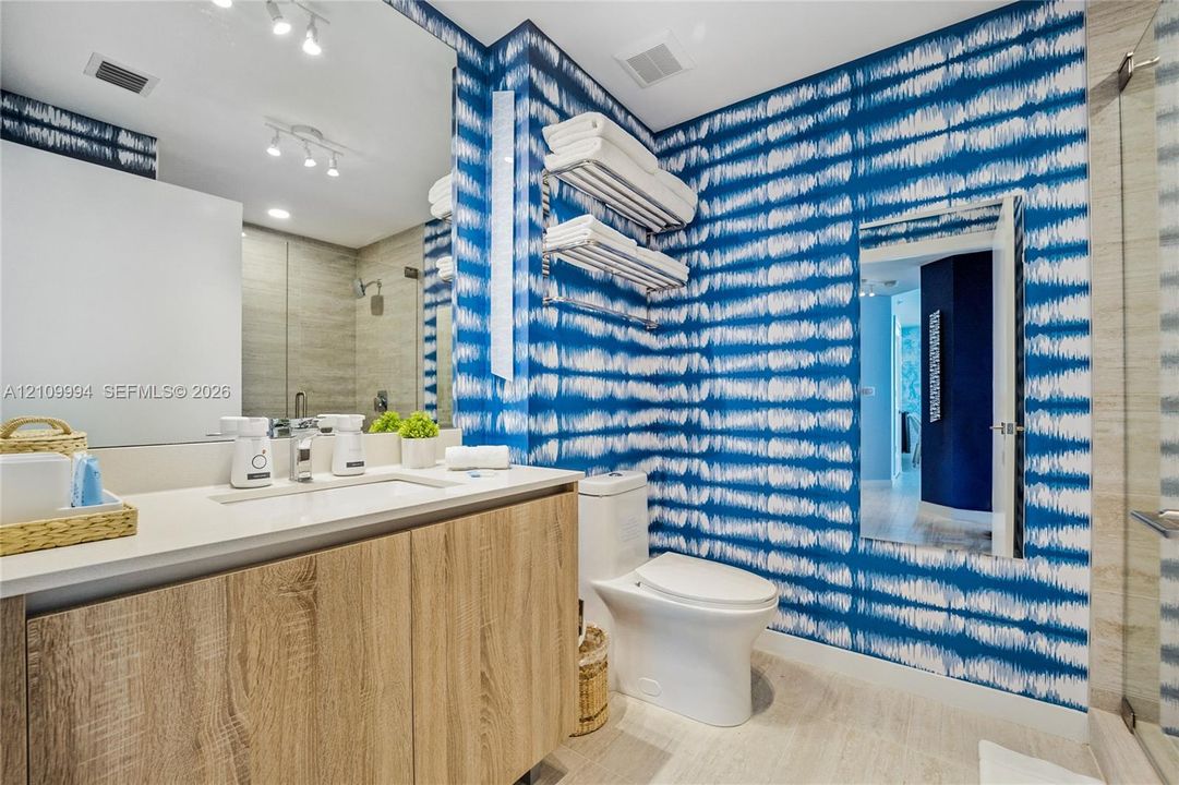 Bathroom featuring a wood-finish floating vanity with a white countertop, wall-to-wall mirror, integrated storage shelving, and a glass-enclosed shower
