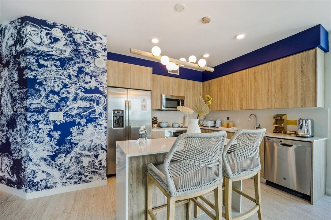 Kitchen featuring wood-finish cabinetry, stainless steel appliances, a waterfall-edge countertop, patterned wall covering, and a multi-bulb linear pendant light