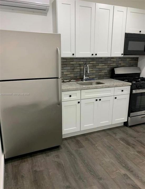 Kitchen featuring white shaker-style cabinetry, stone-finish countertops, brick-style tile backsplash, stainless steel appliances, and wood-finish flooring