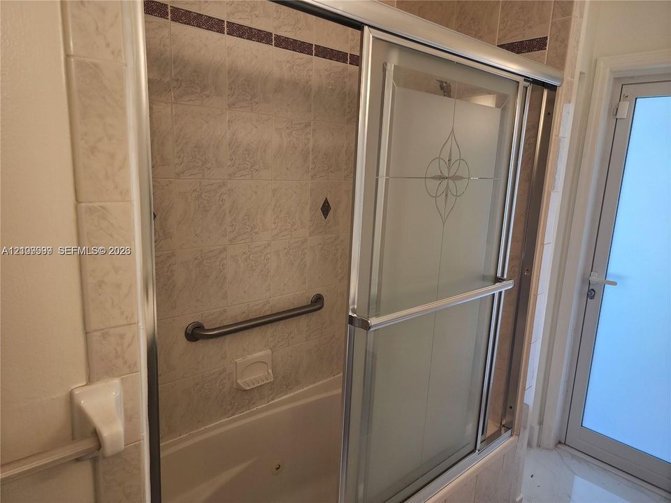 Tub and shower combination featuring a glass enclosure with decorative etching, integrated wall-mounted grab bar, built-in soap dish, and neutral ceramic tile surround with accent detailing