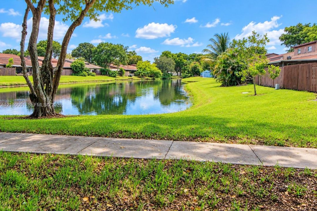 Waterfront grounds featuring a concrete walking path, mature trees, manicured lawn, and perimeter wood fencing
