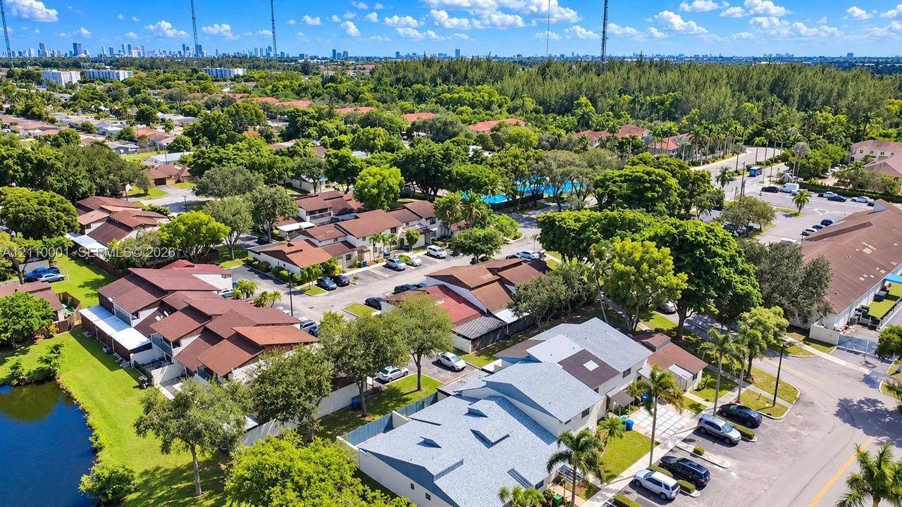 Aerial perspective of a multi-unit residential community featuring brown shingle roofs, paved parking areas, dense mature landscaping, and a water feature