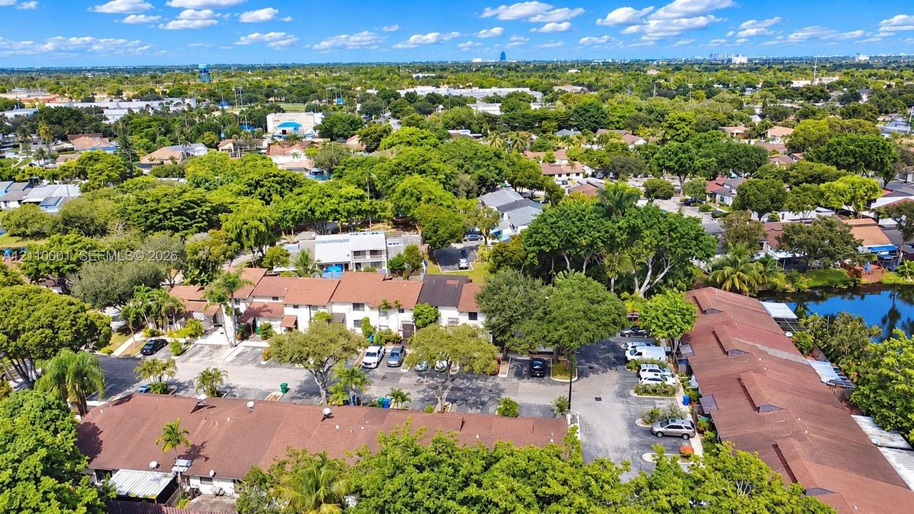 Aerial perspective featuring multiple multi-unit buildings with shingle roofing, paved surface parking, mature canopy trees, and an integrated waterfront pond