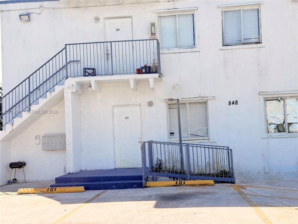 Stucco exterior building featuring a metal staircase, an elevated balcony with metal railings, a ground-level concrete ramp with railings, and designated parking spaces