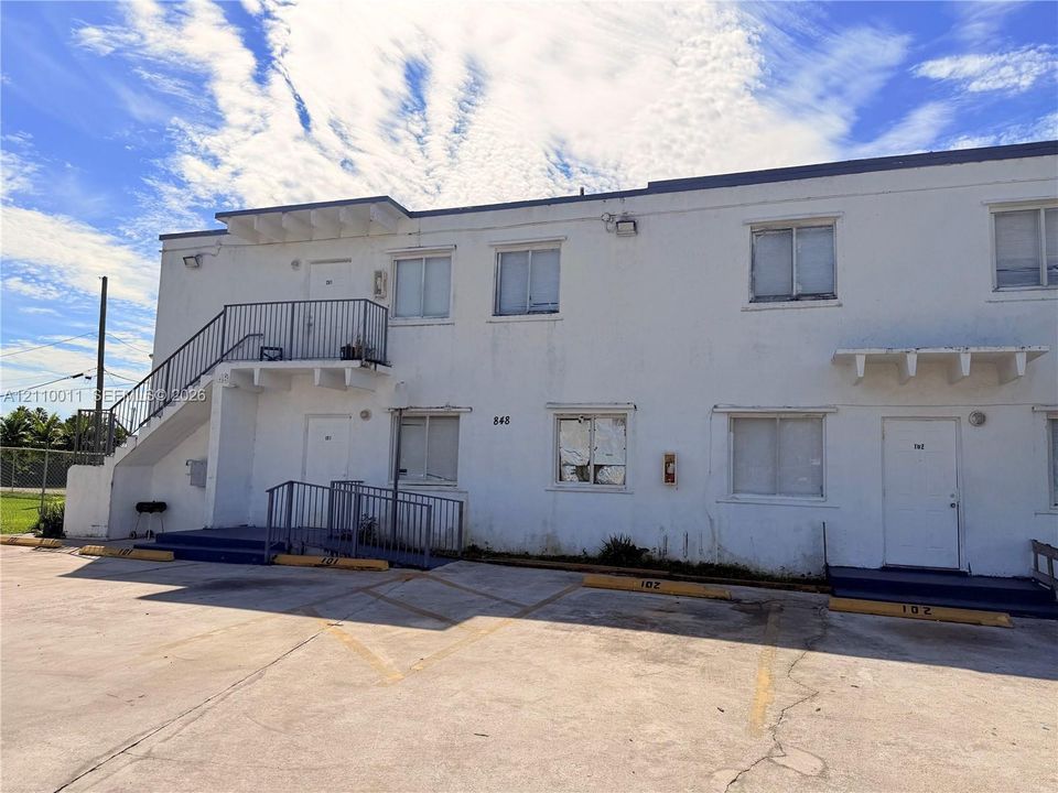 Two-story exterior featuring white stucco finish, external metal staircase, ground-level ramp access, and asphalt-paved parking