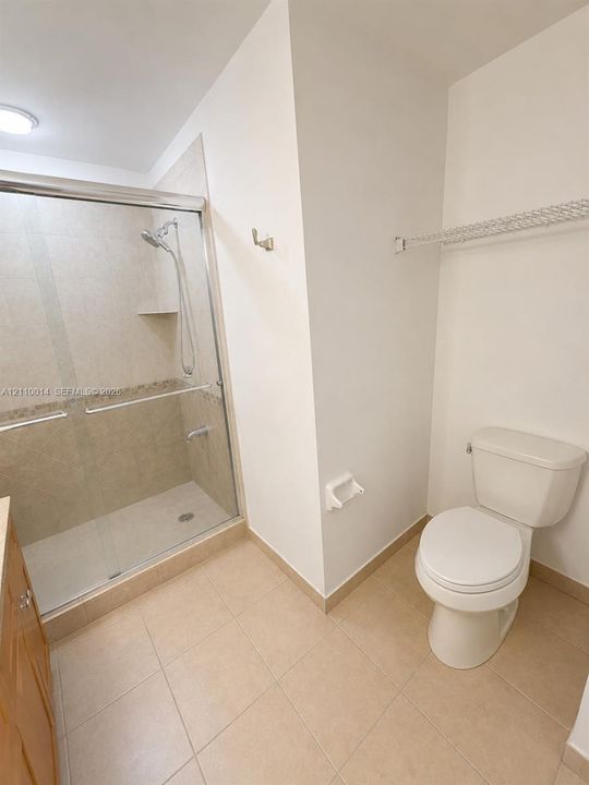 Walk-in shower with a glass enclosure, tile surround, and adjustable handheld shower head, accompanied by tile flooring, a wall-mounted wire storage shelf, and a white porcelain toilet