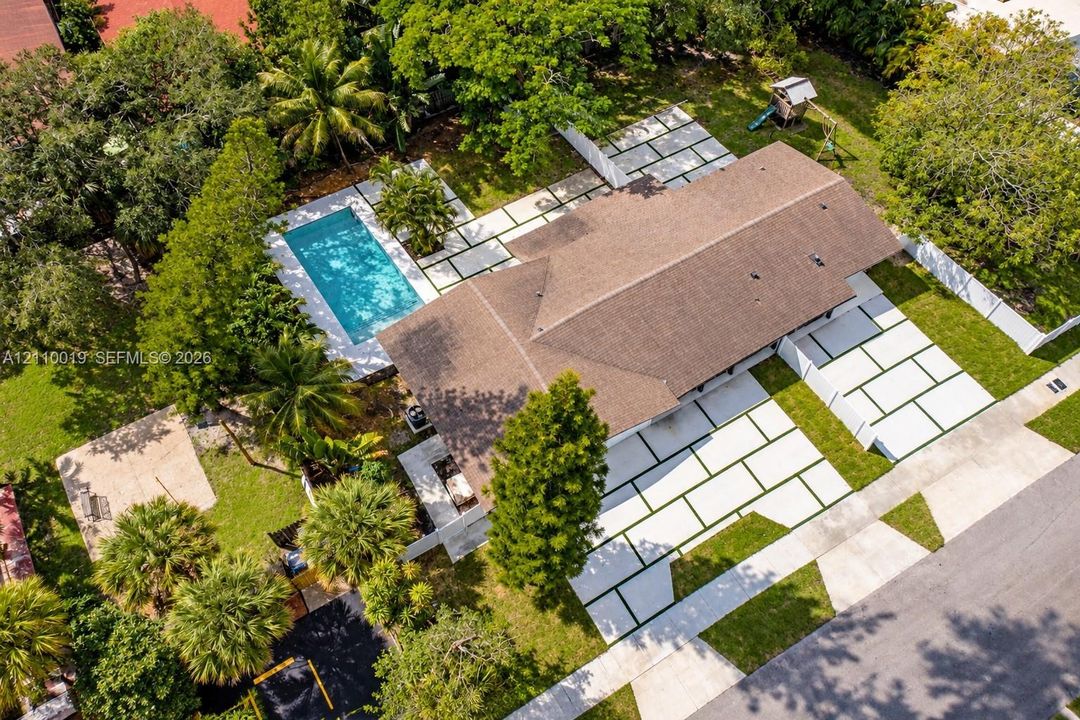 Aerial perspective of a property featuring an in-ground swimming pool, extensive paver hardscaping with geometric inlay, mature landscape, and a fenced perimeter