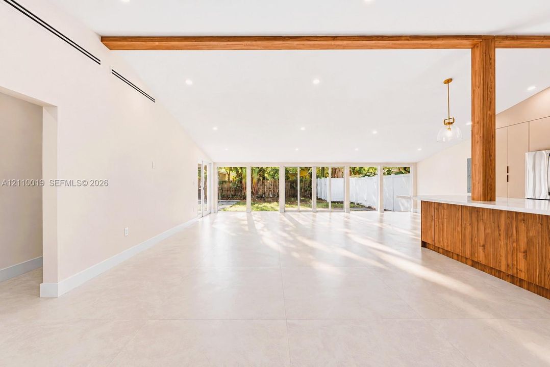 Open-concept interior featuring large-format tile flooring, exposed wood ceiling beams, floor-to-ceiling glass panel windows, and a wood-finish kitchen island with a white countertop