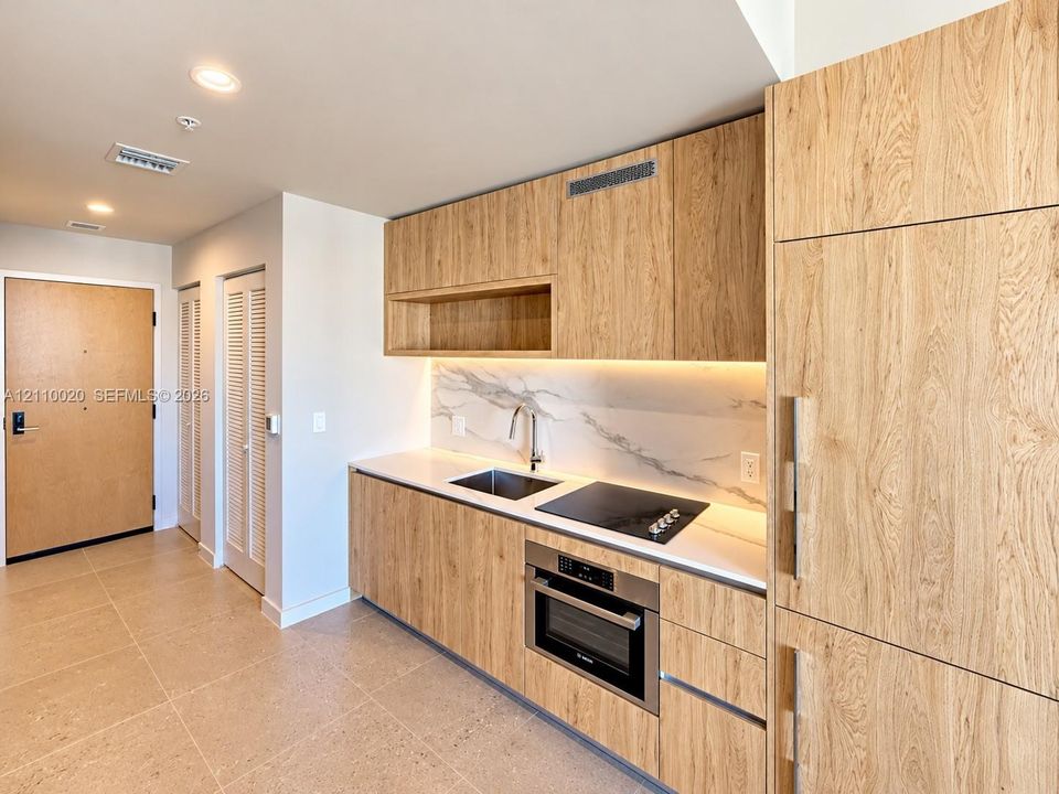 Kitchen area featuring wood-finish cabinetry, solid surface countertops, integrated appliances, stone-patterned backsplash, and recessed lighting