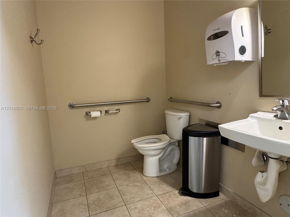 Restroom featuring tiled flooring, a wall-mounted sink with a chrome-finish faucet, an elongated porcelain toilet, and wall-mounted metal support bars