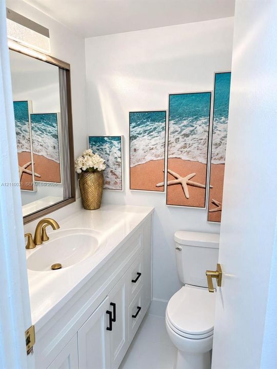 Vanity with white solid-surface countertop and undermount sink, brass-finish widespread faucet, white shaker-style cabinetry with matte black hardware, and rectangular vanity mirror with integrated light fixture