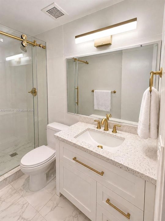 Bathroom featuring a white vanity with speckled countertop, gold-toned hardware, glass-enclosed shower, and large-format tile flooring