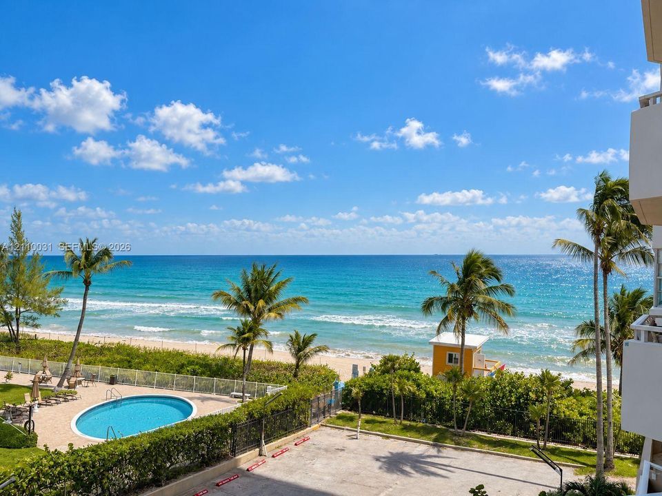 Oceanfront perspective featuring a swimming pool, paved surface parking, perimeter fencing, and mature palm trees