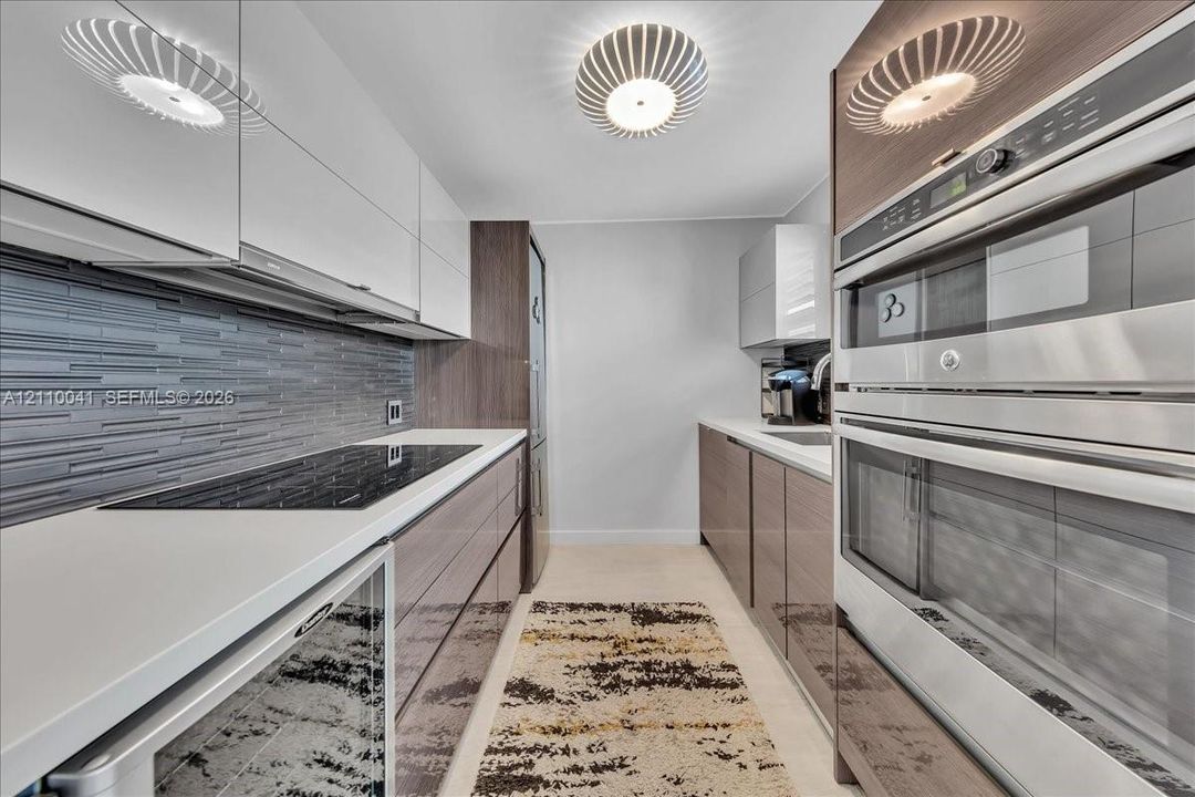 Contemporary kitchen featuring high-gloss cabinetry, stainless steel double wall ovens, a glass cooktop, stone-finish countertops, and a textured tile backsplash