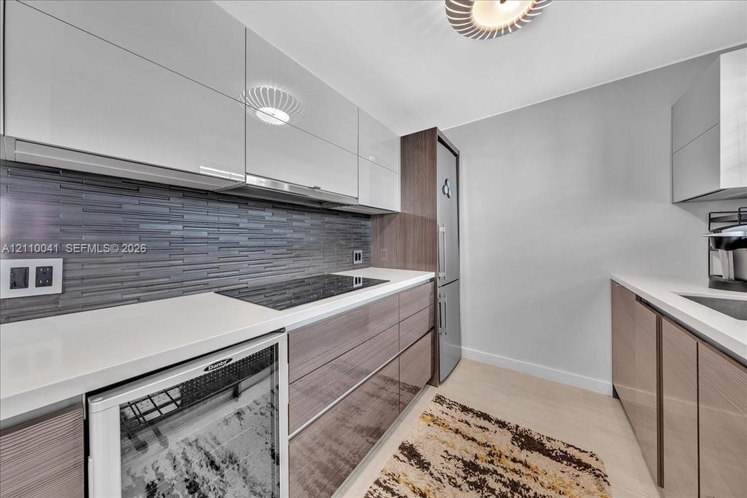 Modern kitchen featuring high-gloss cabinetry, linear tile backsplash, solid-surface countertops, integrated cooktop, and built-in beverage refrigerator