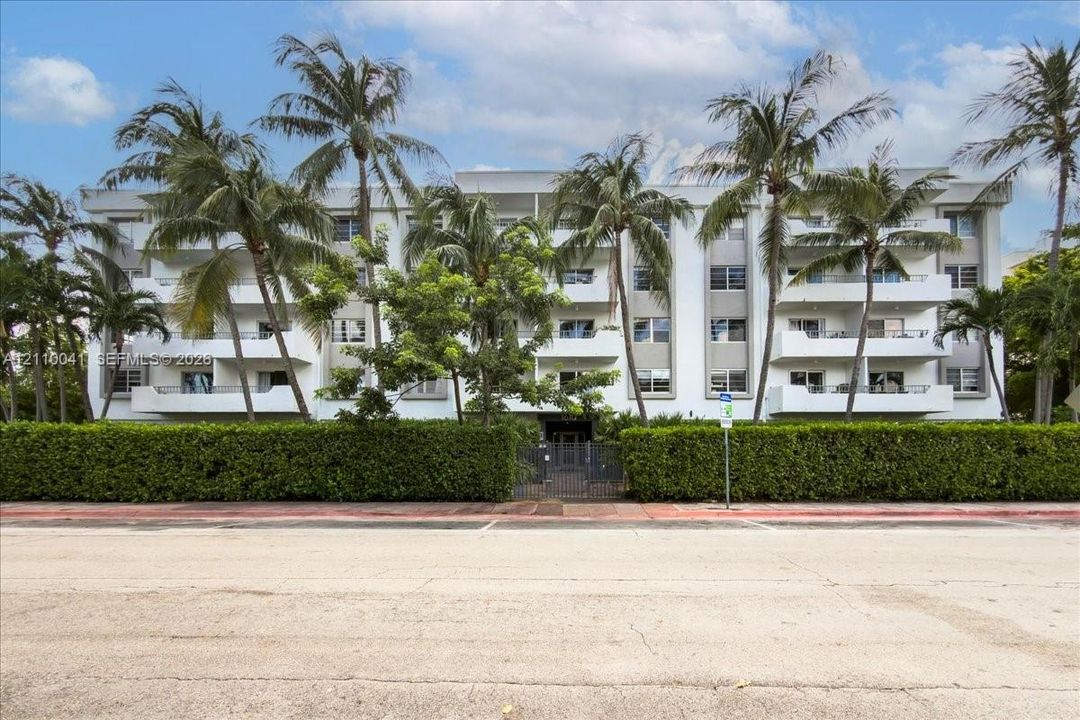 Multi-story residential building featuring private balconies, vertical architectural pillars, and mature palm tree landscaping set behind a manicured hedge