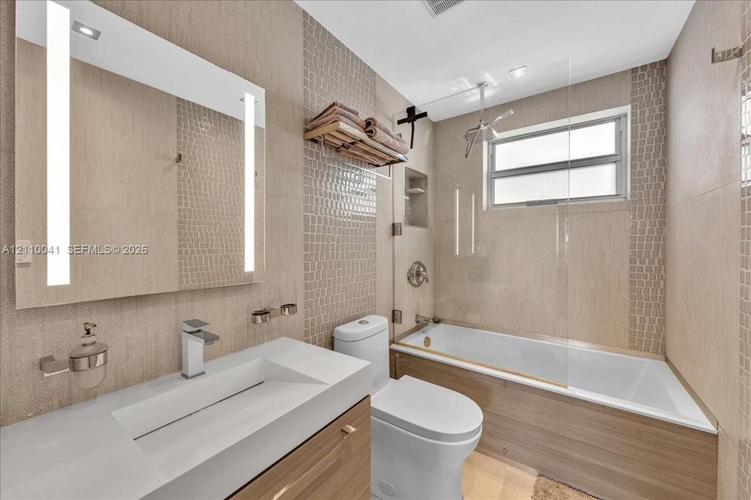 Bathroom featuring a wood-finish vanity with a white rectangular vessel sink, a wall-mounted LED-backlit mirror, a glass-enclosed shower and tub combination, and neutral textured tile wall accents
