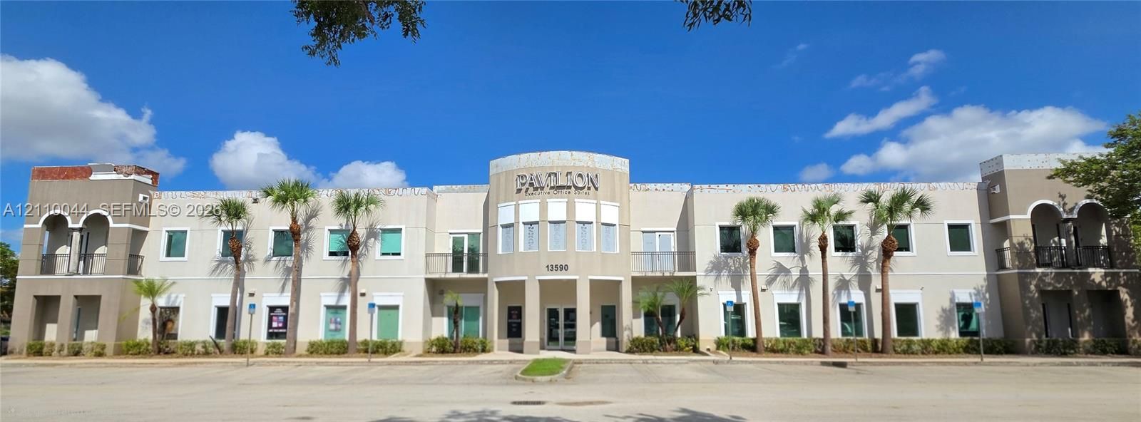 Two-story commercial building exterior featuring a stucco facade, arched architectural details, second-floor balconies, and mature palm landscaping. Ca you spot the unit's balcony to the right of the front entrance???