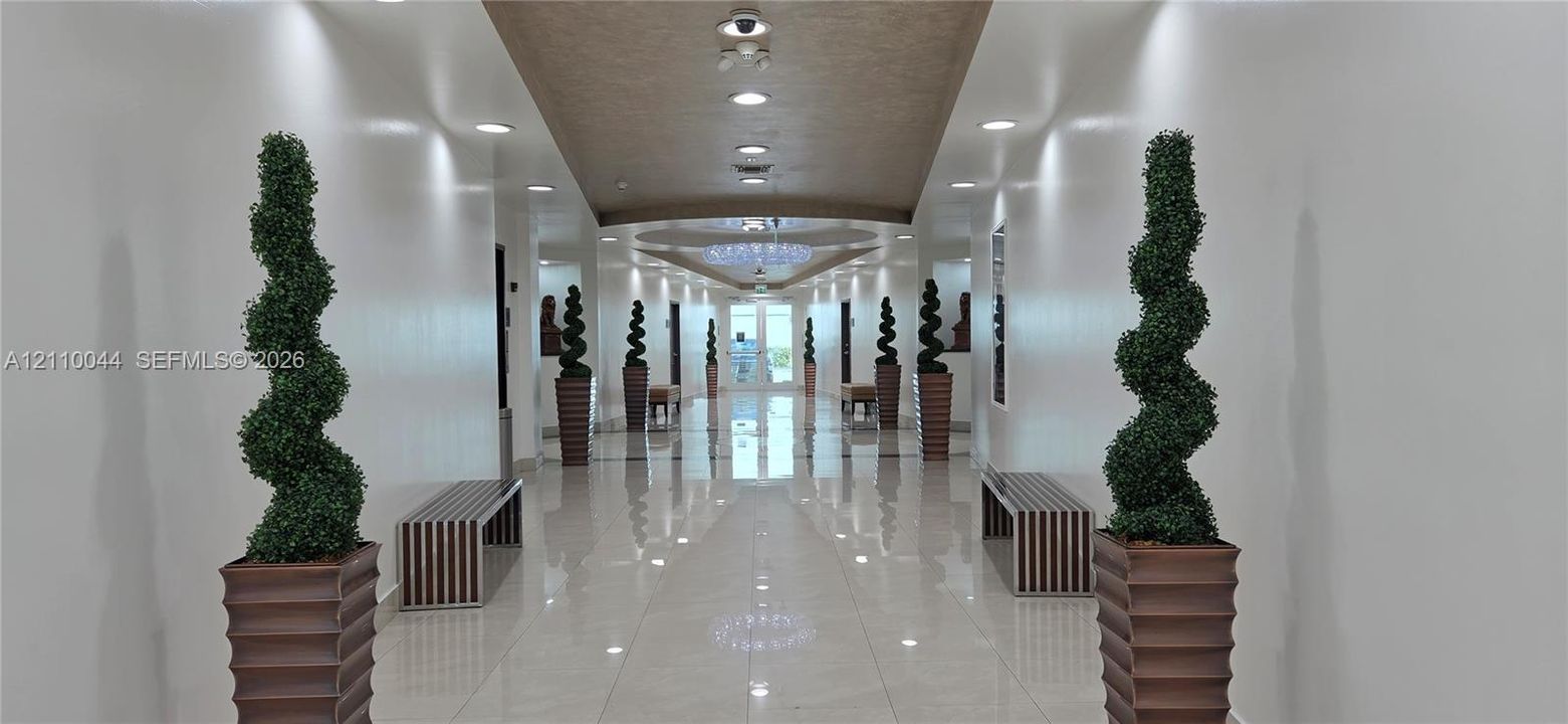 Wide hallway featuring high-gloss tile flooring, a recessed ceiling with a central crystal-style chandelier, and integrated recessed lighting