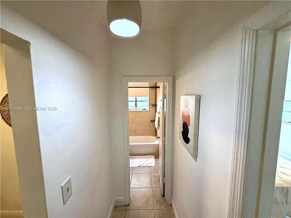 Hallway featuring neutral-toned walls, tile flooring, a semi-flush mount light fixture, and a doorway opening into a bathroom containing a porcelain tub and tile surround