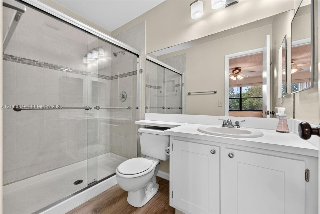 Full-width vanity with an integrated sink, glass-enclosed shower with mosaic tile inlay, wood-finish flooring, and wall-to-wall mirror