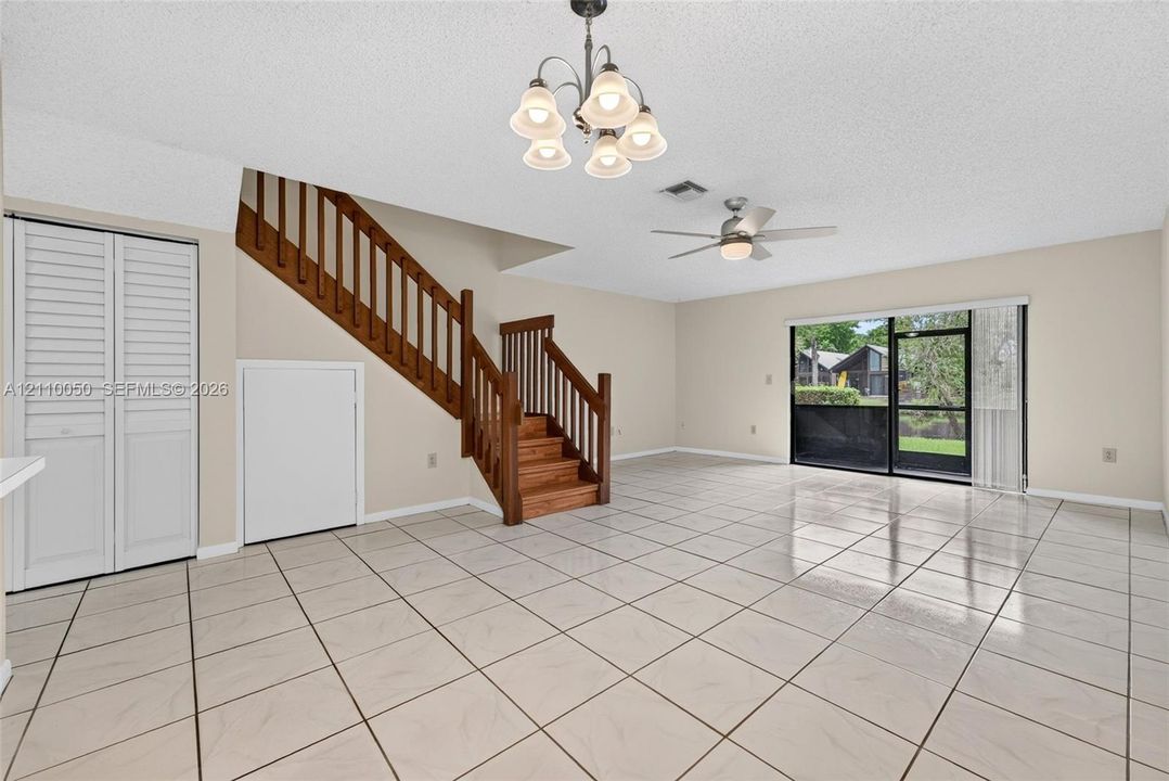 Open-concept living space featuring ceramic tile flooring, a wooden staircase with integrated railing, sliding glass doors, a multi-arm chandelier, and a ceiling fan