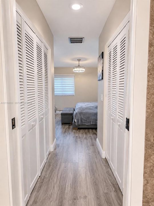 Hallway featuring dual sets of white louvered closet doors, wood-finish flooring, and a vanity area equipped with a mirror, three-light fixture, and stone-finish cabinetry