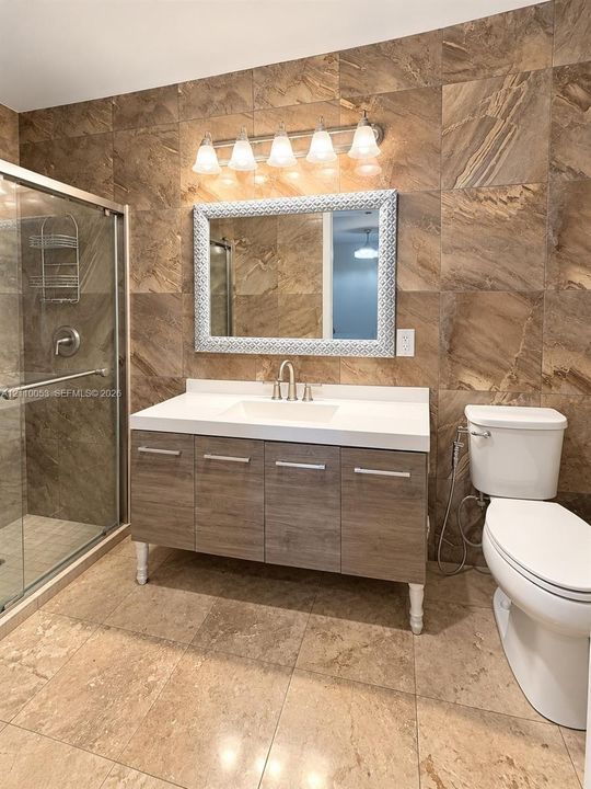 Bathroom featuring floor-to-ceiling stone-tile walls, a glass-enclosed shower with sliding doors, a vanity with a white integrated basin, and wood-finish cabinetry