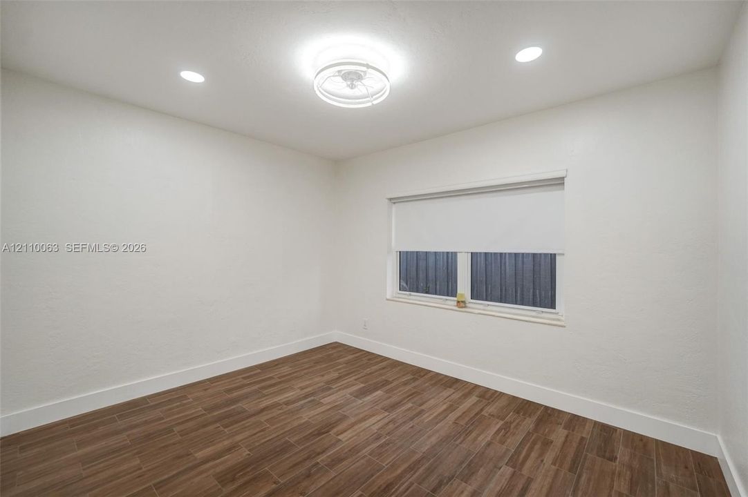 Neutral-toned interior featuring wood-finish flooring, recessed lighting, a central flush-mount ceiling fixture, and a single-pane window with a fitted roller shade