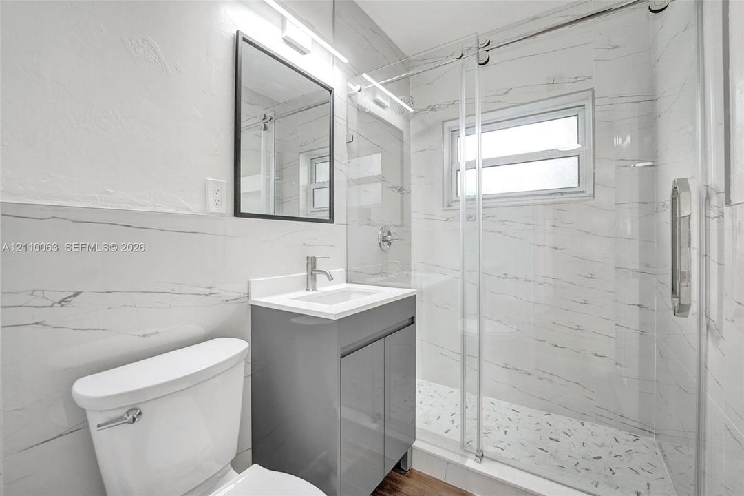 Bathroom featuring full-height large-format wall tile, glass-enclosed shower, stone-finish vanity with rectangular basin, and black-framed wall mirror