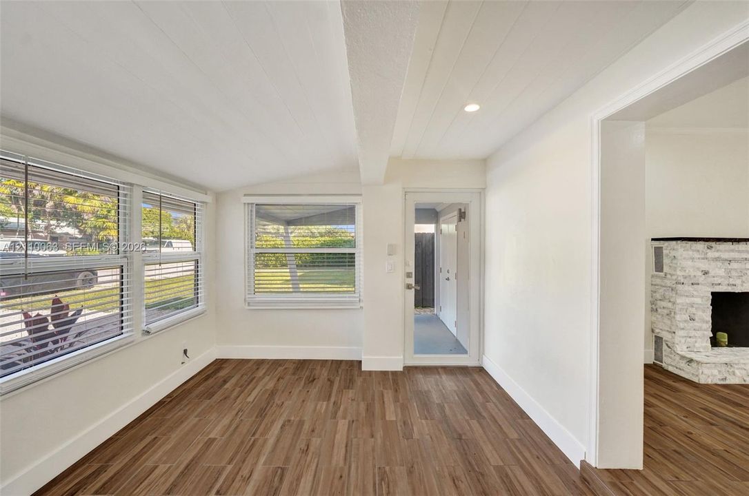 Wood-finish flooring, white plank-style ceiling, multi-pane windows, glass-panel exterior door, and a whitewashed brick fireplace