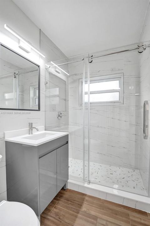 Modern vanity with a rectangular sink, walk-in shower with sliding glass doors, floor-to-ceiling wall tile, wood-finish flooring, and wall-mounted light fixture