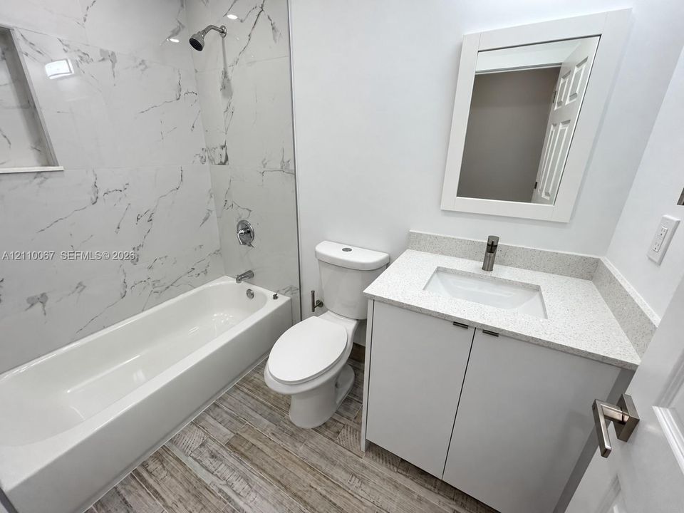 Bathroom featuring large-format tile tub surround, wood-finish flooring, white vanity with stone-composite countertop, undermount sink, and wall-mounted mirror