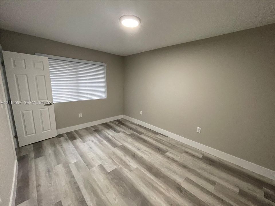 Neutral-toned interior space featuring wood-finish flooring, a six-panel door, and a horizontal window with adjustable blinds