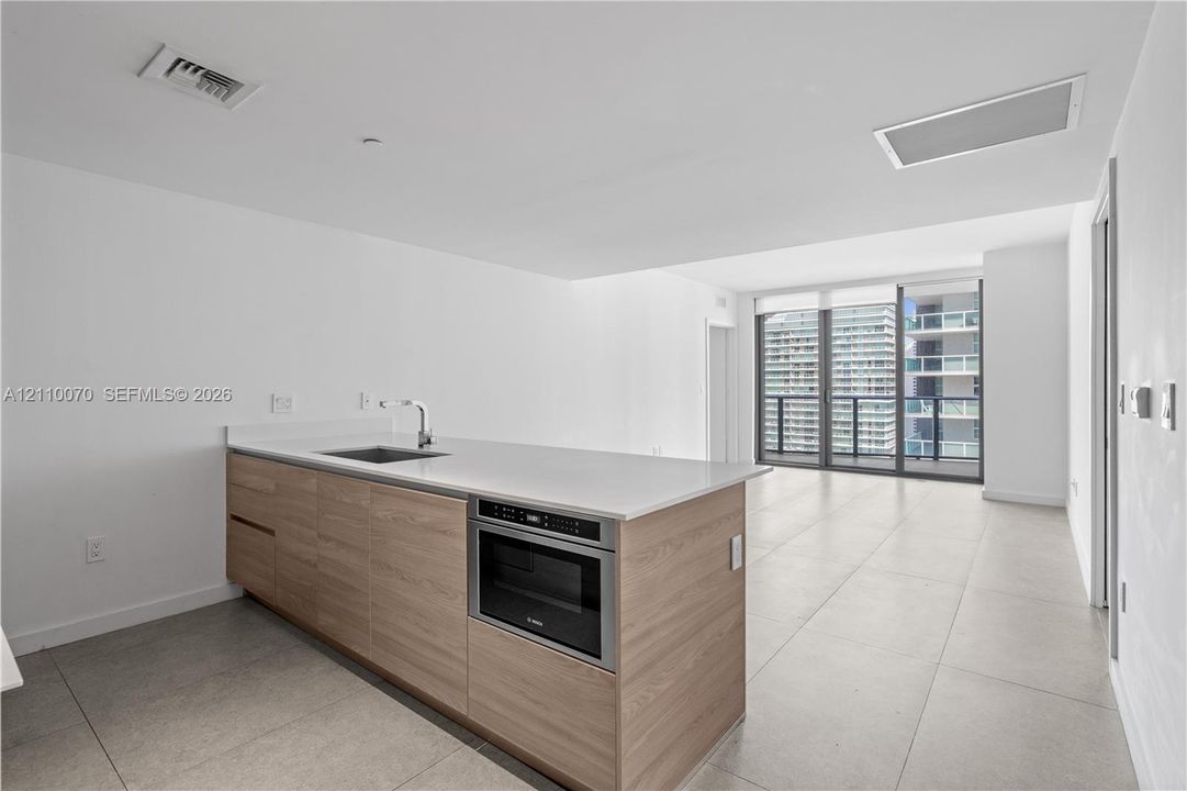 Kitchen island featuring wood-finish cabinetry, solid surface countertop, stainless steel sink with high-arc faucet, and built-in microwave, complemented by large-format tile flooring and floor-to-ceiling glass doors