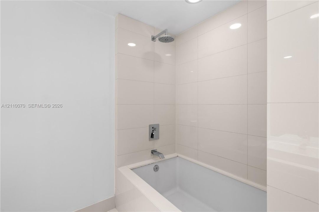 Full-height neutral wall tiling, integrated bathtub, chrome shower head, chrome wall-mounted faucet, and recessed ceiling lighting