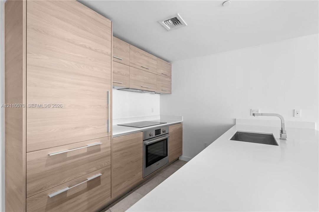 Modern kitchen featuring flat-panel wood-finish cabinetry, white solid-surface countertops, built-in stainless steel appliances, and an undermount sink with a gooseneck faucet